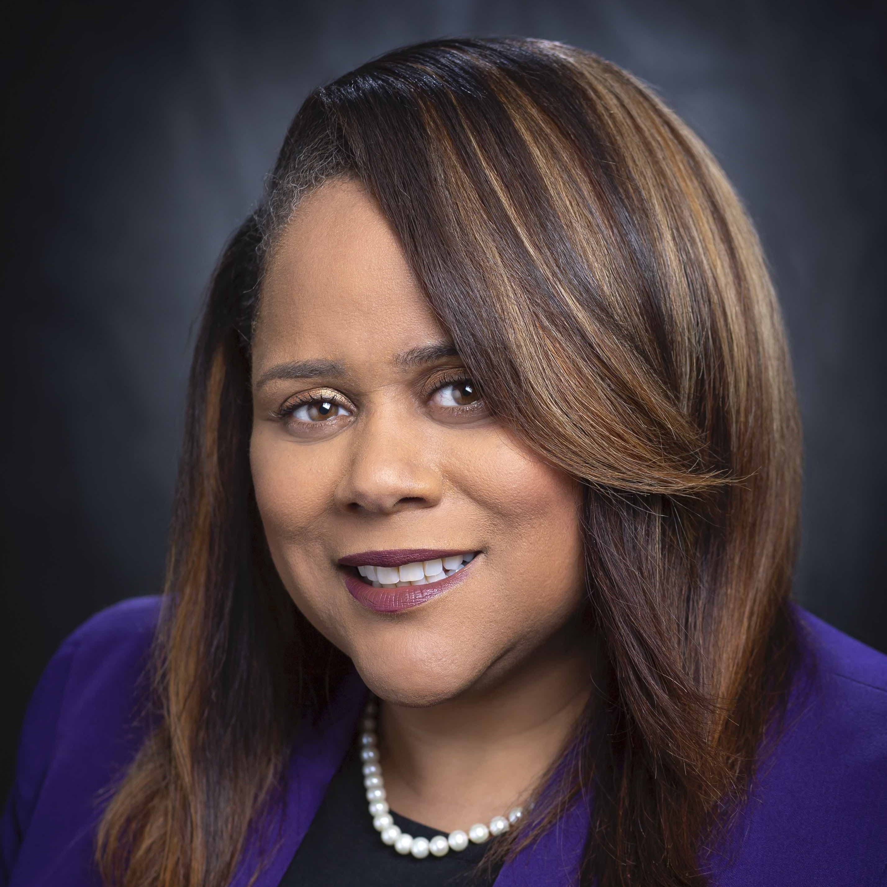 Keena Arbuthnot headshot is in a purple jacket on a dark background.